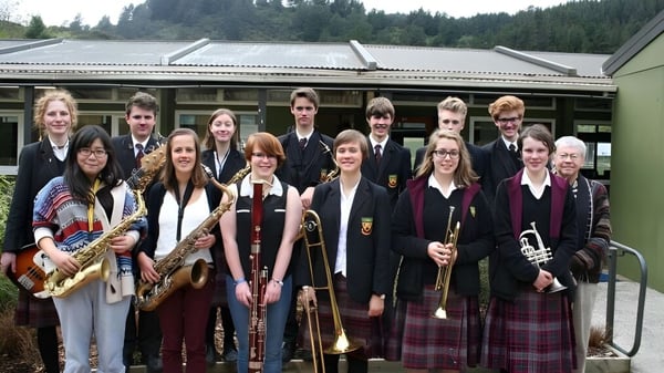 Eine Gruppe Schüler der Logan Park High School mit Musikinstrumenten steht vor einem Gebäude vor bewaldetem Hintergrund.