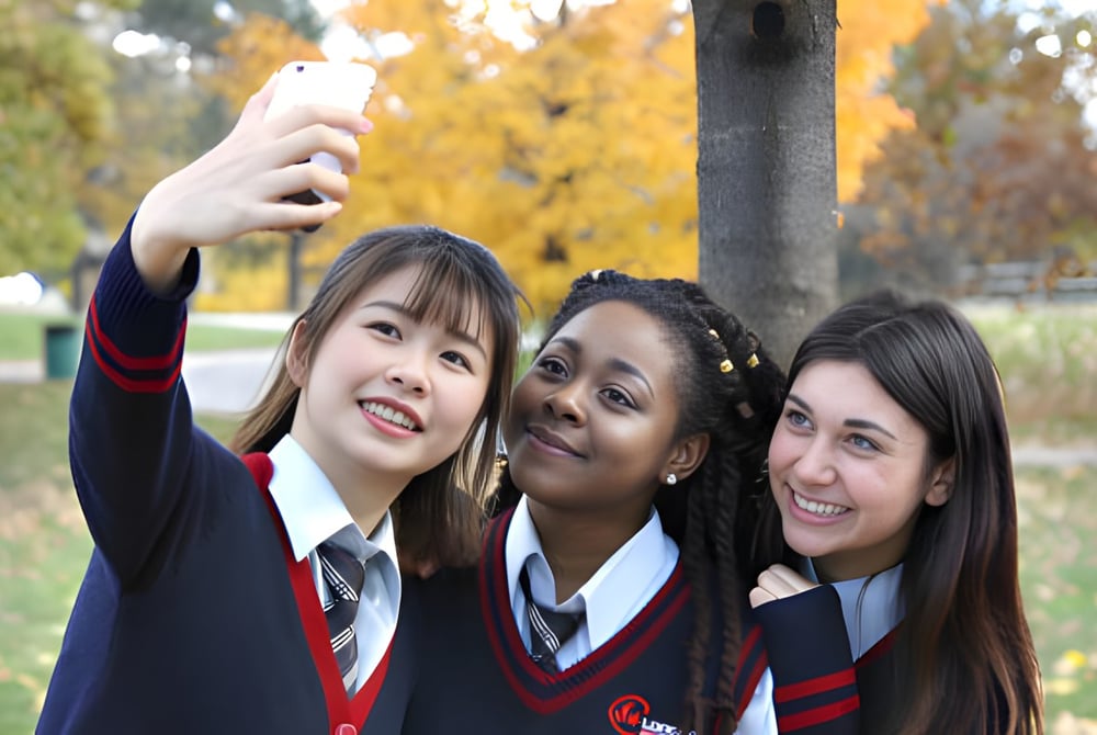 Drei Schülerinnen der London International Academy machen ein Selfie im Park mit herbstlicher Laubkulisse.