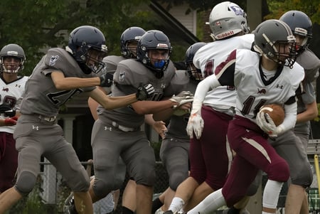 Eine Gruppe von Football-Spielern der London South Collegiate Institute versammelt sich auf dem Spielfeld mit Bäumen und Gebäuden im Hintergrund.