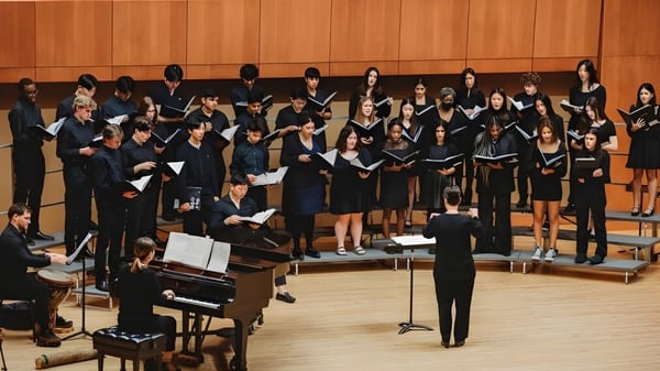 Der Chor der Loomis Chaffee School steht in schwarzen Gewändern auf der Bühne vor einer Holzpaneelwand mit einem Klavier im Vordergrund.