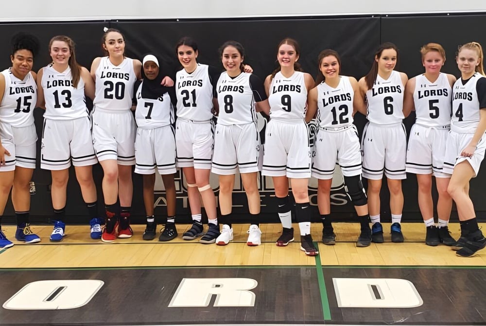 Eine Gruppe junger Basketballspielerinnen in weißen Trikots steht auf dem Basketballfeld der Lord Beaverbrook High School.