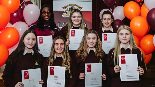Eine Gruppe junger Absolventinnen des Loreto College Cavan steht mit Zeugnissen vor Luftballons und dem Schullogo.