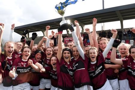 Schülerinnen des Loreto College Cavan feiern ihren Sieg mit einem Pokal in einer Sportarena.
