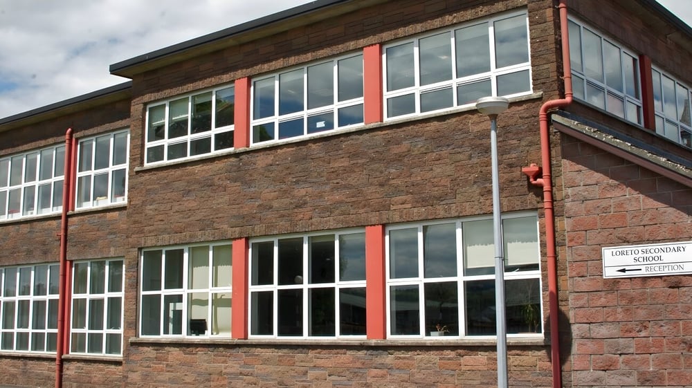 Das mehrstöckige Backsteingebäude der Loreto Secondary School in Cork ist mit großen Fenstern und roten Akzenten unter bewölktem Himmel zu sehen.
