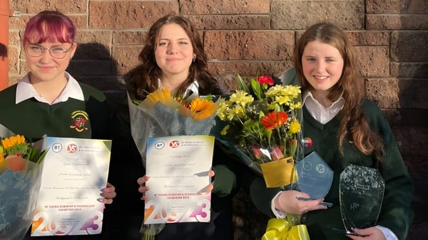 Drei Schülerinnen der Loreto Secondary School in Cork halten Zertifikate und Blumen vor einer Backsteinwand.