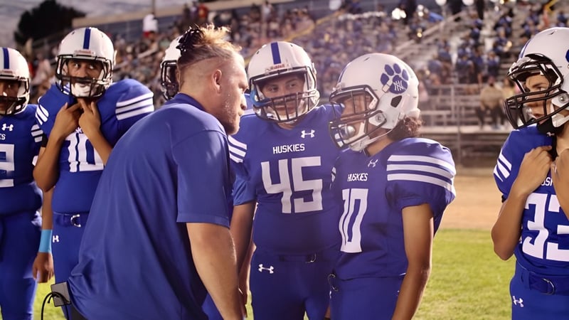 Eine Gruppe von Footballspielern in blauen Uniformen steht auf dem Spielfeld des Los Angeles Unified School District mit Zuschauern im Hintergrund.