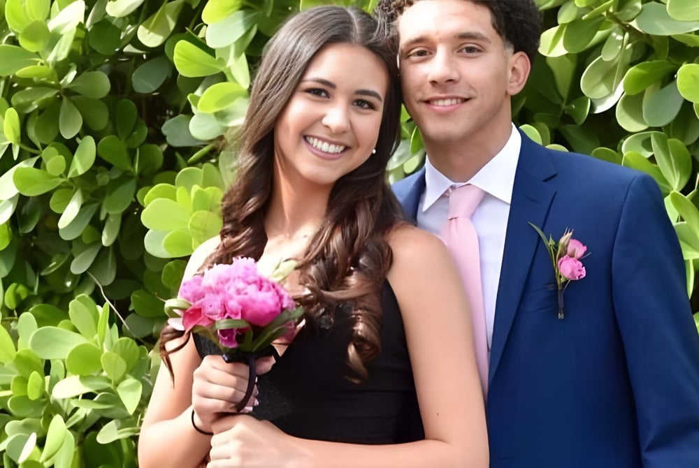 Ein Paar steht lächelnd vor einem grünen Hintergrund mit Blumen auf dem Campus der Lourdes Academy.
