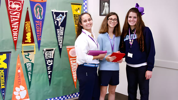 Drei Schülerinnen stehen in einem Raum mit bunten Schulpennants und Bannern auf den Wänden der Lourdes Academy.