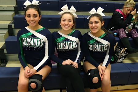 Drei Schülerinnen der Lowell Catholic High School posieren gemeinsam in Cheerleader-Uniformen in einer Sporthalle.