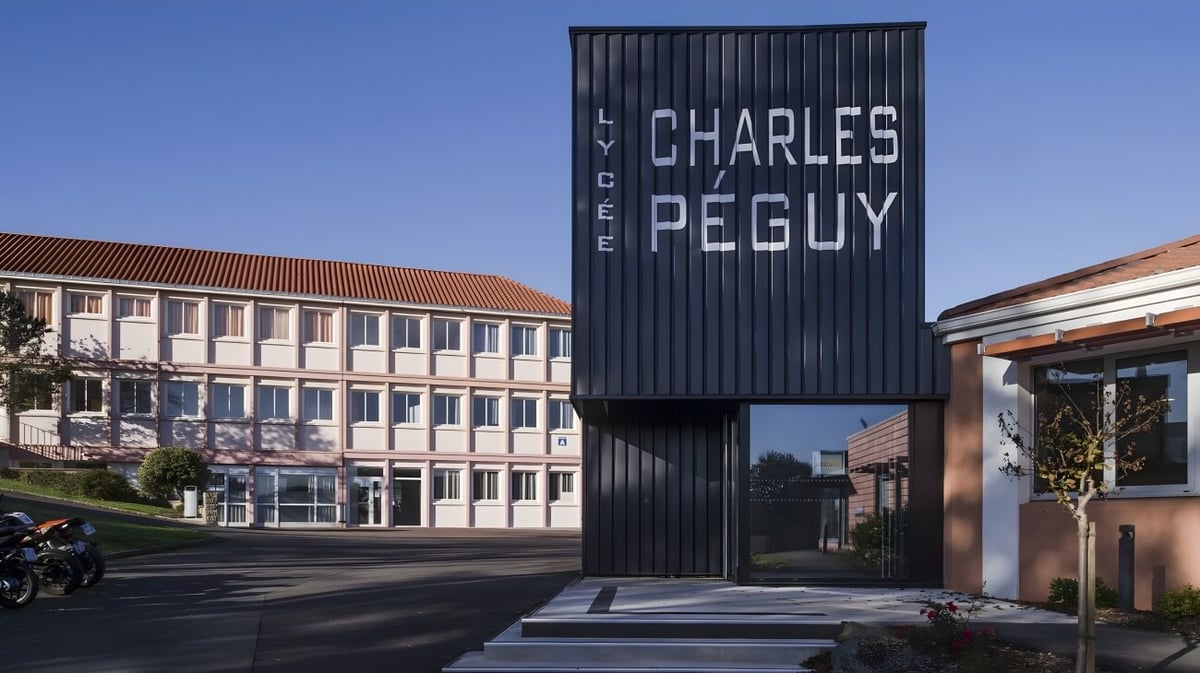 Ein großes schwarzes Schild mit der Aufschrift Lycée Charles Péguy vor einem mehrstöckigen Schulgebäude.