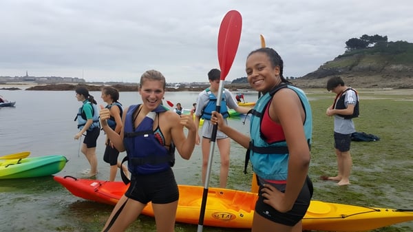 Eine Gruppe von Schülerinnen und Schülern des Lycée Jean Brito steht in bunter Kajakausrüstung am Strand vor einer Küstenlandschaft.