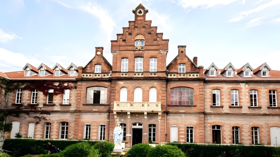 Das Gebäude des Lycée Notre Dame de Bon Secours mit einem markanten Treppengiebel und Bogenfenstern ist von üppigem Grün umgeben.