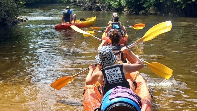 Schüler des Lycée St Elme paddeln mit bunten Kajaks einen Fluss entlang, umgeben von grüner Vegetation.