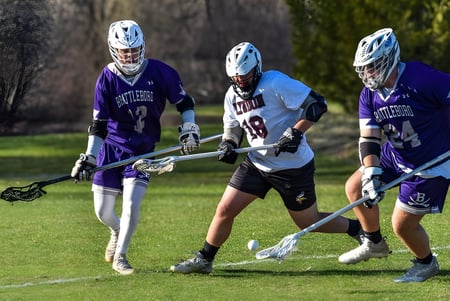 Drei Spieler in Lacrosse-Uniformen beim Wettkampf auf dem Sportfeld des Lyndon Institute bei Nacht.