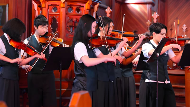 Eine Gruppe Musiker in formeller Kleidung tritt auf der Bühne des Lynfield College vor rotem und goldenem Hintergrund auf.