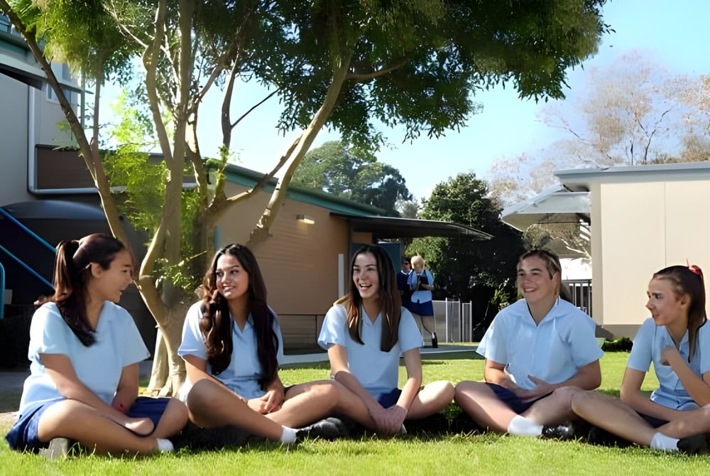 Eine Gruppe junger Schülerinnen sitzt auf dem Gras auf dem Gelände des Mackellar Girls Campus, umgeben von Bäumen und Gebäuden.