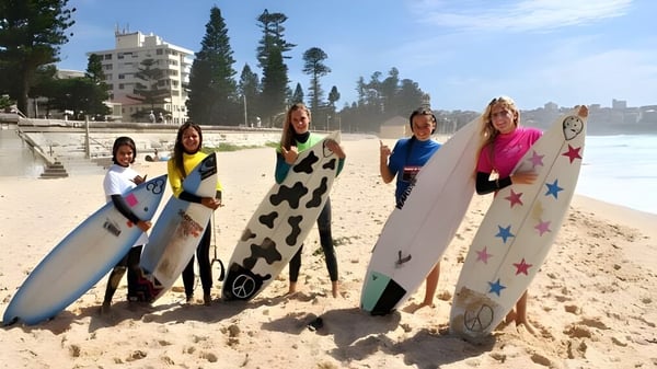 Schülerinnen der Mackellar Girls Campus stehen mit bunten Surfboards am Strand vor Gebäuden und Bäumen.