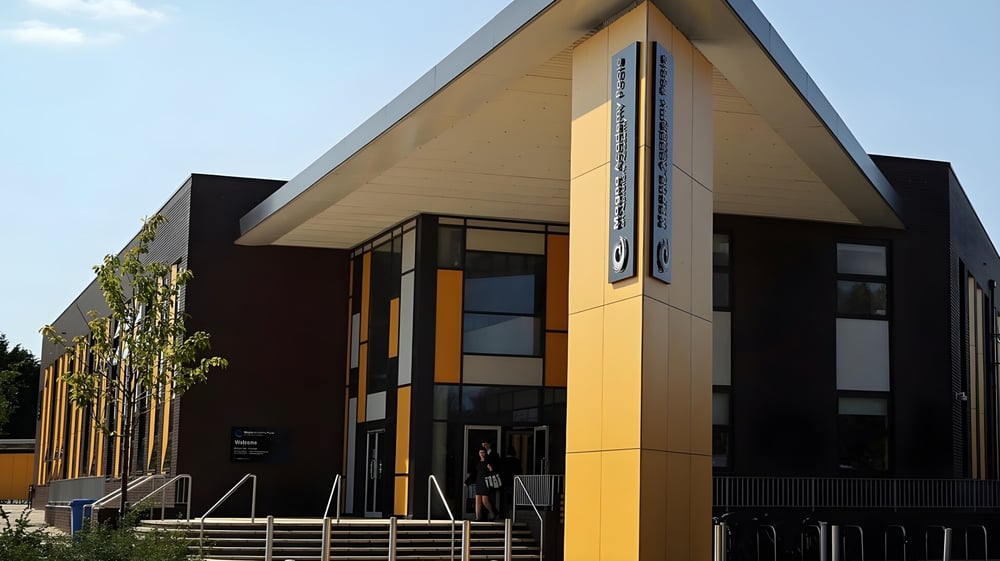 Das moderne Gebäude der Magna Academy mit schwarzen und gelben Fassadenpaneelen ist von einem gepflasterten Bereich und Vegetation umgeben.