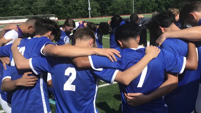 Eine Gruppe Schüler der Magna Academy stehen im Kreis auf einem Feld und umarmen sich vor einem Spiel.