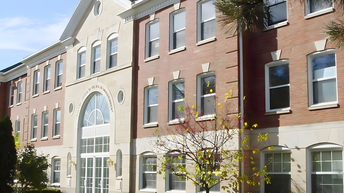 Das mehrstöckige Backsteingebäude der Maharishi School mit bogenförmigen Fenstern und einem Baum im Vordergrund bei klarem Himmel.