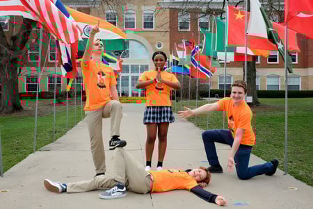 Schüler der Maharishi School stehen in orangefarbenen Shirts vor einem Campusgebäude mit internationalen Flaggen.