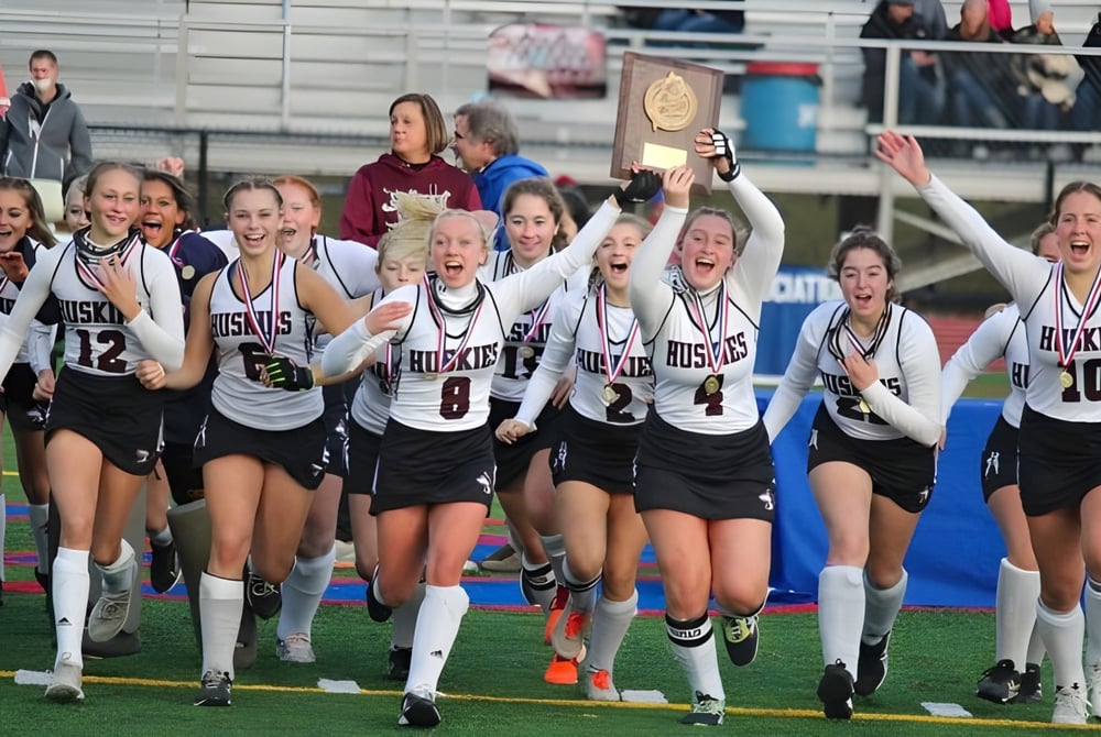 Eine Gruppe junger Athletinnen des Maine Central Institute feiert auf dem Sportplatz den Gewinn eines Pokals.