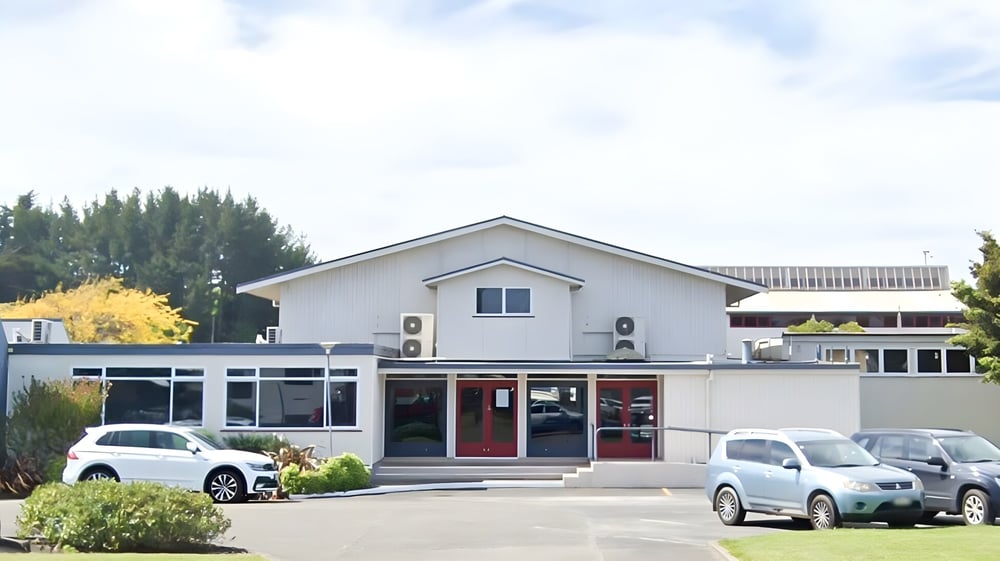 Das moderne weiße Gebäude mit schrägem Dach auf dem Campus des Manawatu College ist von geparkten Fahrzeugen und Bäumen umgeben.