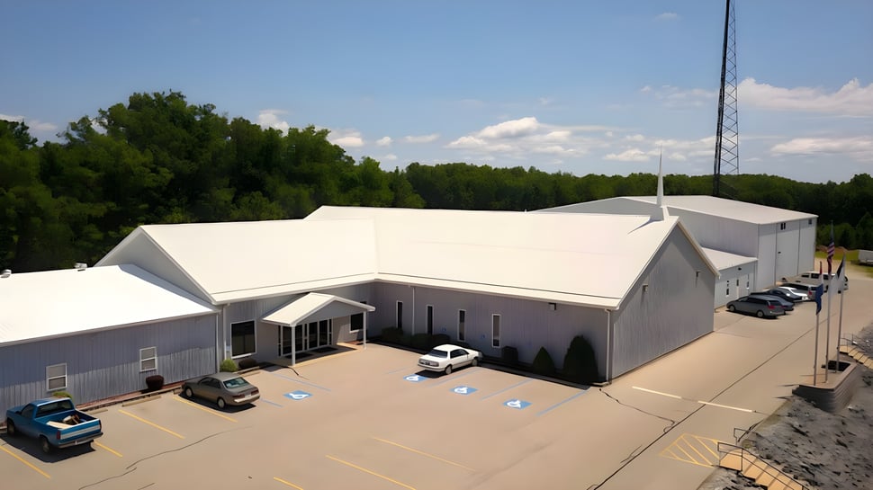 Das weiße Industriegebäude der Maranatha Baptist Academy steht in einem bewaldeten Gebiet mit geparkten Fahrzeugen im Vordergrund.