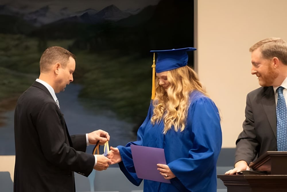 Eine Schülerin in blauer Abschlussrobe der Maranatha Baptist Academy schüttelt einem Mann im Anzug die Hand vor einer Berg- und Waldkulisse.