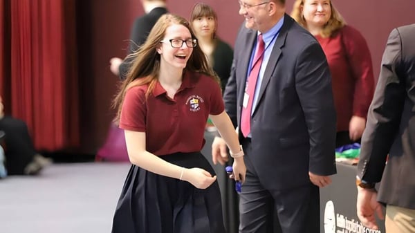 Eine Schülerin der Maranatha Baptist Academy lächelt mit Brille vor Erwachsenen in formeller Kleidung.
