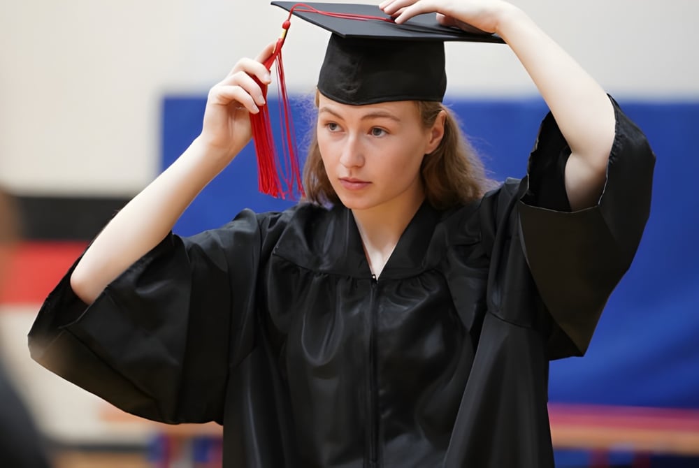 Eine Absolventin der École secondaire publique Marc-Garneau steht im Raum vor blauem Hintergrund mit ihrem schwarzen Abschlussmantel und -mütze in den Händen.