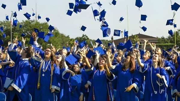 Eine große Gruppe von Absolventinnen und Absolventen der Marion County Public Schools wirft die blauen Abschlussmützen in die Luft vor einem Baumhintergrund.