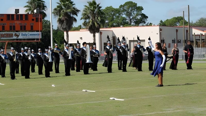 Eine Gruppe uniformierter Schüler der Marion County Public Schools steht in Formation auf einem Rasenfeld mit Palmen und Gebäuden im Hintergrund.
