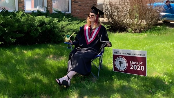 Eine Absolventin in Talaren steht vor einem Schild mit der Aufschrift Class of 2020 auf dem Campus der Marion M. Graham Collegiate.
