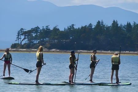 Schüler der Mark R. Isfeld Secondary School beim Paddleboarding auf einem See umgeben von Wald und Bergen.
