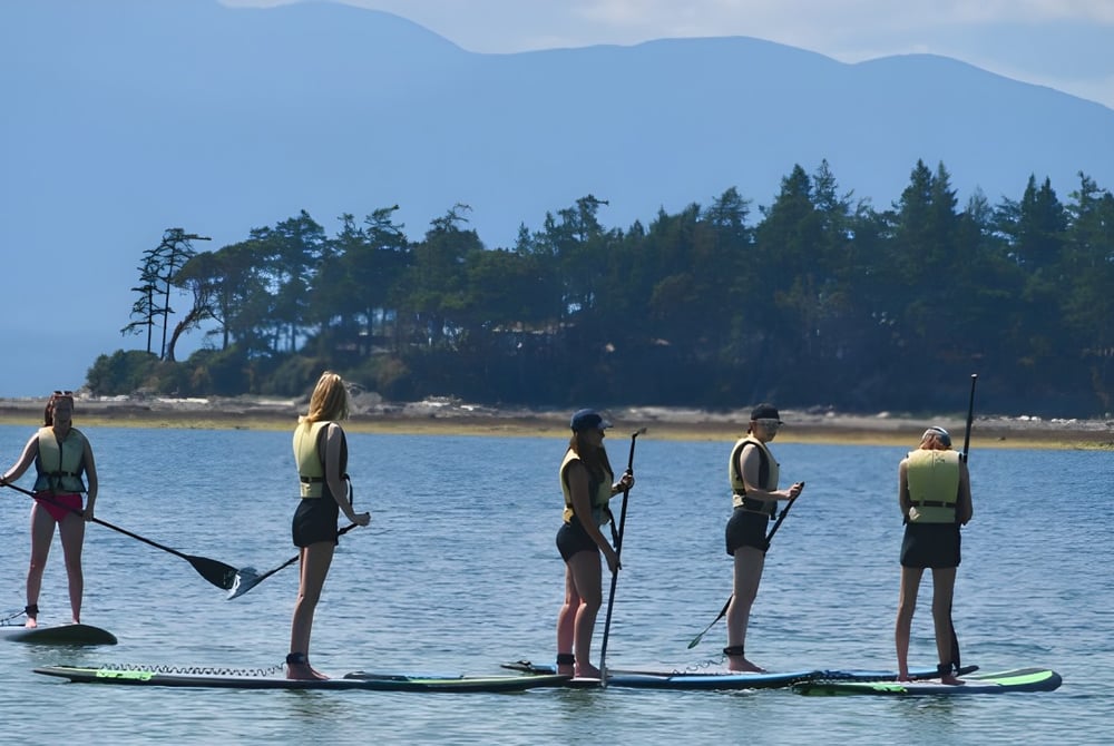 Schüler der Mark R. Isfeld Secondary School beim Paddleboarding auf einem See umgeben von Wald und Bergen.