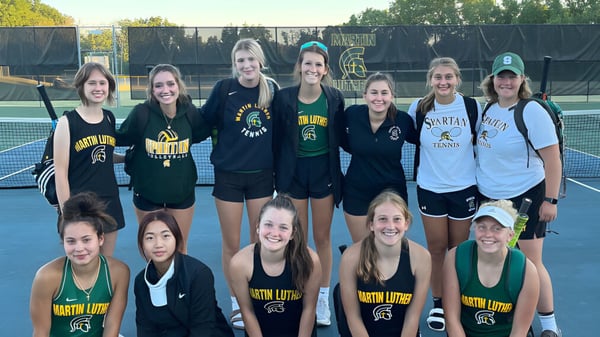 Eine Gruppe Schülerinnen der Martin Luther High School posiert gemeinsam auf dem Tennisplatz vor einem Zaun mit Bäumen im Hintergrund.