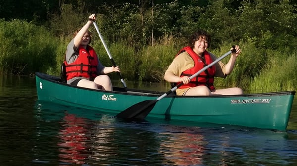 Zwei Schüler der Marvelwood School paddeln mit Schwimmwesten ein grünes Kanu auf einem von Vegetation umgebenen Gewässer.