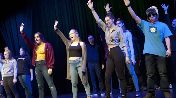 Schüler der Marymount Academy International stehen auf einer Bühne mit erhobenen Armen vor dunklem Hintergrund.