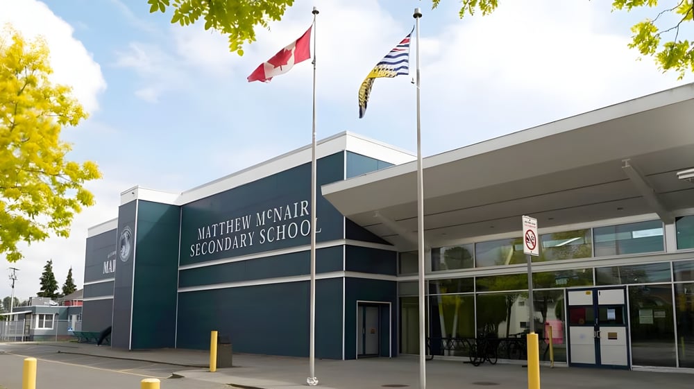Das moderne mehrstöckige Schulgebäude der Matthew McNair Secondary School ist von Bäumen umgeben und zeigt mehrere Fahnen im Außenbereich.
