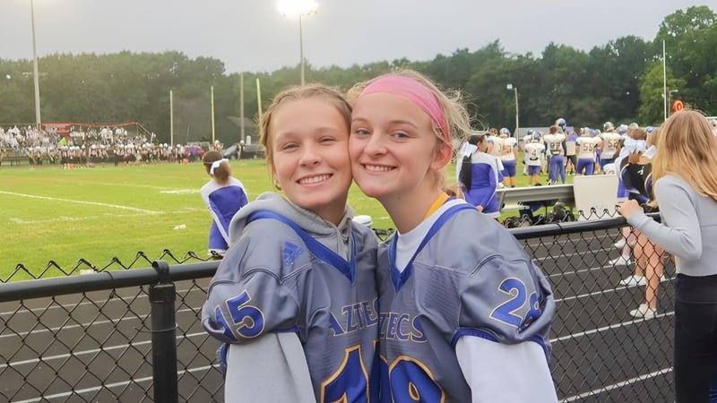 Zwei junge Sportlerinnen stehen lächelnd auf dem Sportfeld der Maynard High School mit Zuschauern im Hintergrund.