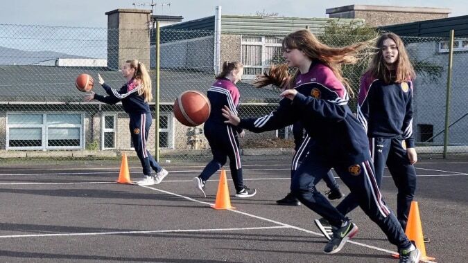 Eine Gruppe Schülerinnen spielt Basketball auf dem Außenspielfeld des McEgan College mit Gebäuden im Hintergrund.