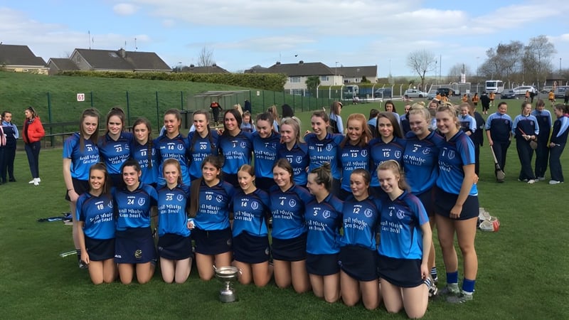 Eine Gruppe junger Athletinnen der Meán Scoil Mhuire Longford posiert mit einer Trophäe auf einem Rasenfeld vor Wohnhäusern.