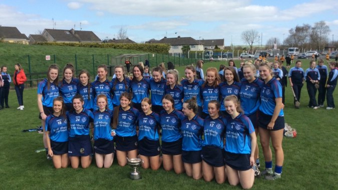 Schülerinnen der Meán Scoil Mhuire in blauen Trikots posieren mit einem Pokal auf dem Sportplatz.