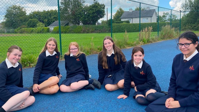 Eine Gruppe junger Schülerinnen der Meán-Scoil Mhuire Gan Smál sitzt auf einer blauen Fläche im Pausenhof mit grüner Wiese und Zaun im Hintergrund.