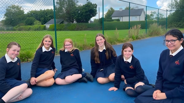 Schülerinnen der Meán-Scoil Mhuire Gan Smál sitzen in Uniform auf einer blauen Gummifläche im Außenbereich der Schule.