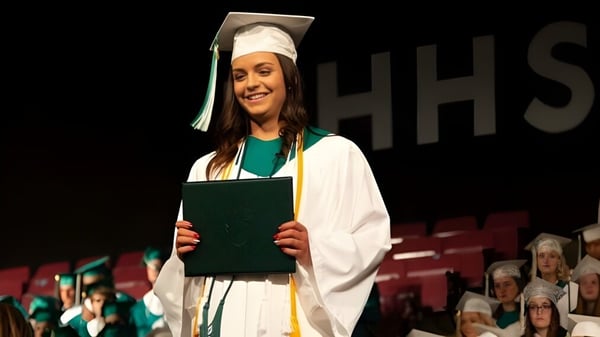 Eine lächelnde Absolventin der Medicine Hat High School hält auf der Bühne ihre Urkunde vor dem Hintergrund mit den Buchstaben HHS.