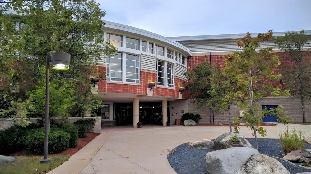 Das moderne mehrstöckige Gebäude der Medway High School mit gebogener Fassade und umgeben von Bäumen zeigt den Eingangsbereich mit gepflastertem Weg.