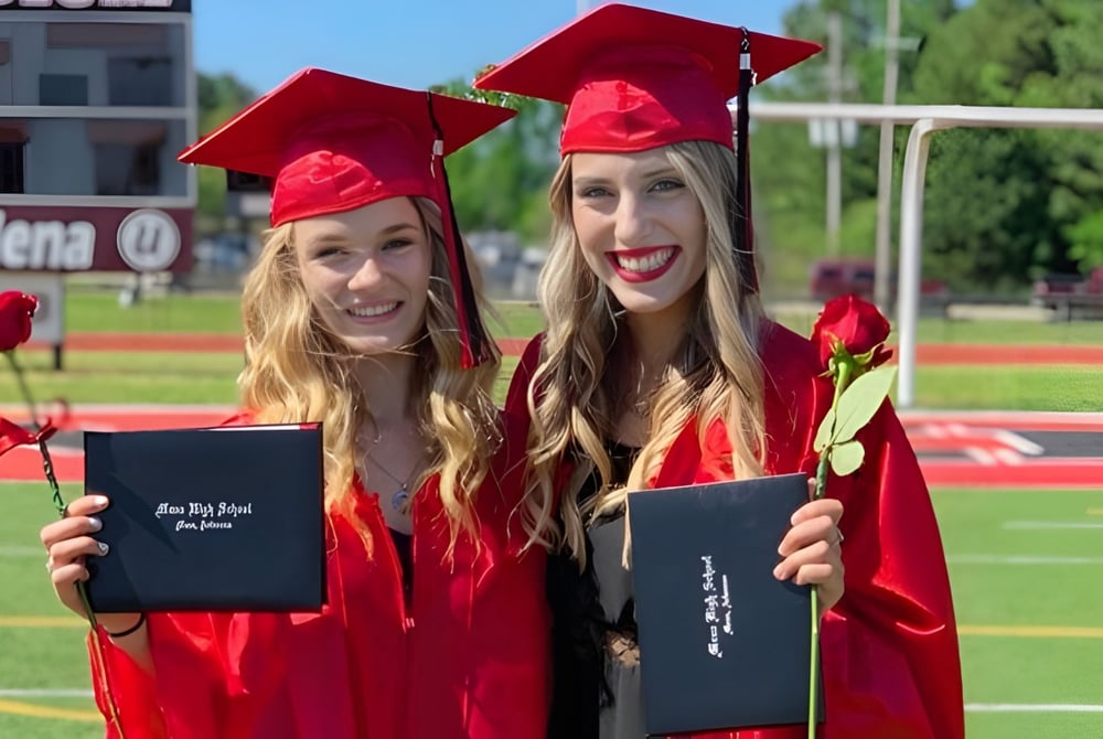 Zwei Absolventinnen der Mena High School stehen auf dem Sportplatz und halten ihre Diplome.