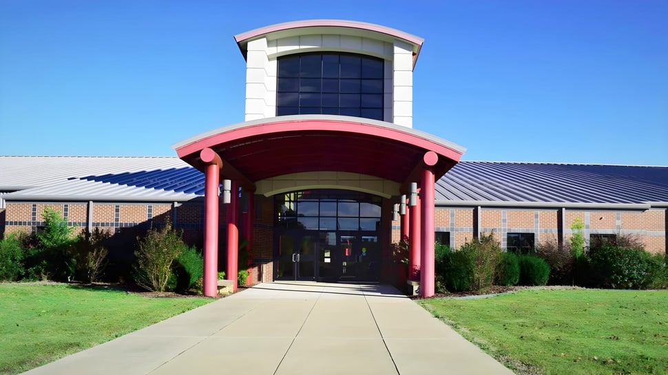 Das moderne mehrstöckige Gebäude der Mena High School mit gläsernem Eingang und rotem Vordach steht auf einer Rasenfläche bei klarem Himmel.